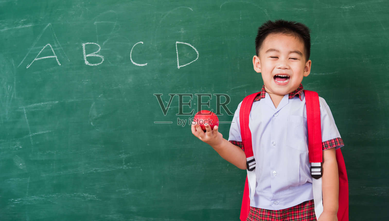 幼儿园的小男孩穿着校服，背着书包，微笑着拿着红苹果照片摄影图片