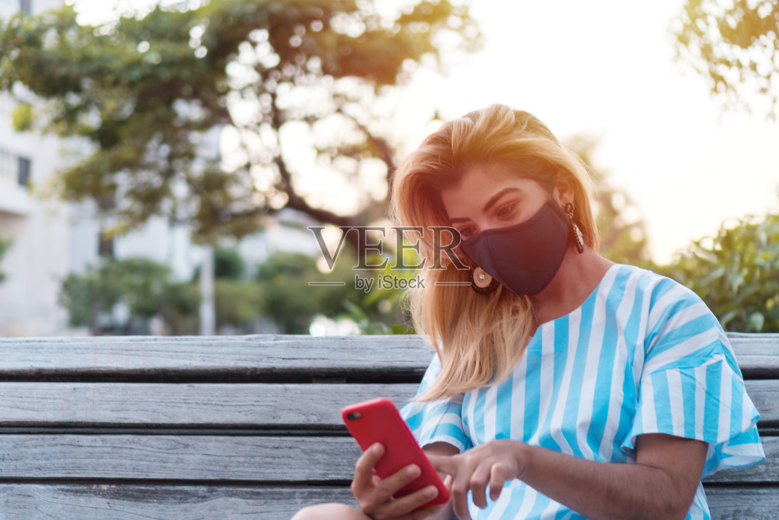 一个带着保护面具和手机的年轻女子的户外肖像。坐在公园里拿着手机的女孩照片摄影图片