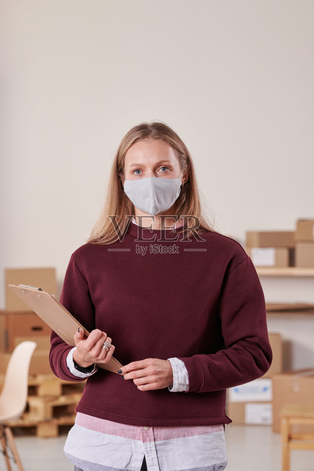 年轻的仓库妇女的肖像戴面具和毛衣站在剪贴板对着纸箱在仓库照片摄影图片