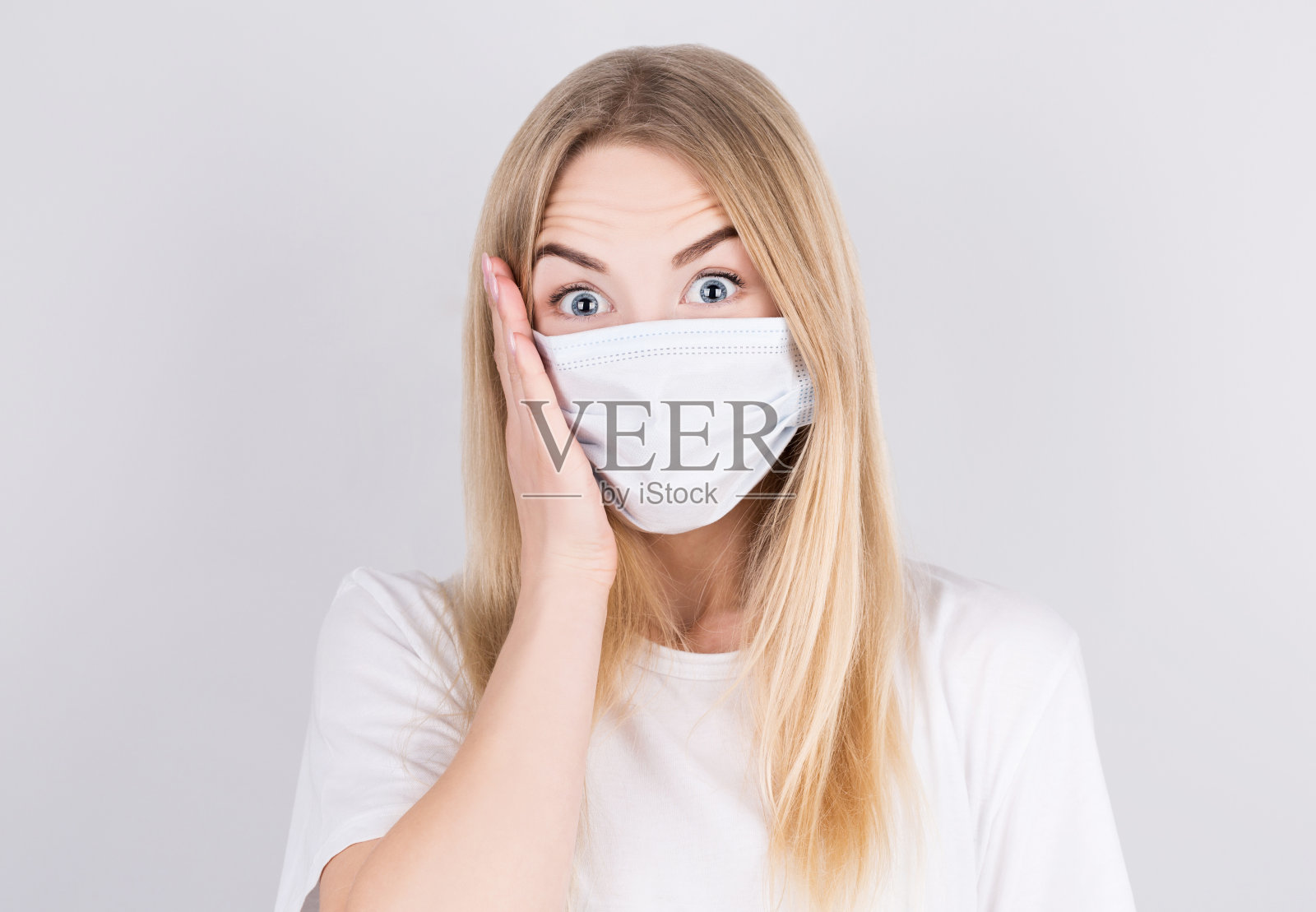 美丽的白人年轻女子在白色t恤与一次性面罩惊讶和看着相机与他的手到他的脸。对病毒和感染的保护。照片摄影图片