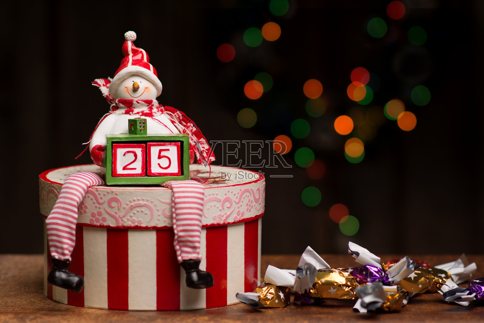一个雪人坐在一个条纹剪纸盒上，拿着一个小盒子，里面有两个立方体组成数字25。旁边放着一堆客厅里的糖果。照片摄影图片