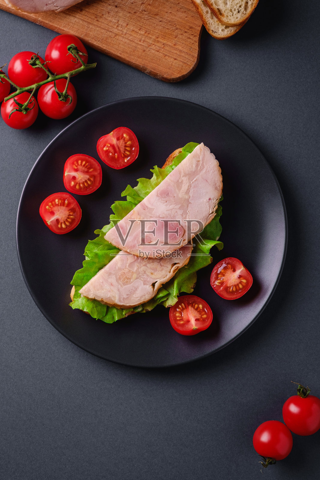 夹有火鸡火腿肉，绿色沙拉和新鲜樱桃番茄片的三明治放在黑色的盘子里，靠近砧板上的食材，灰色的最小背景，俯视图照片摄影图片