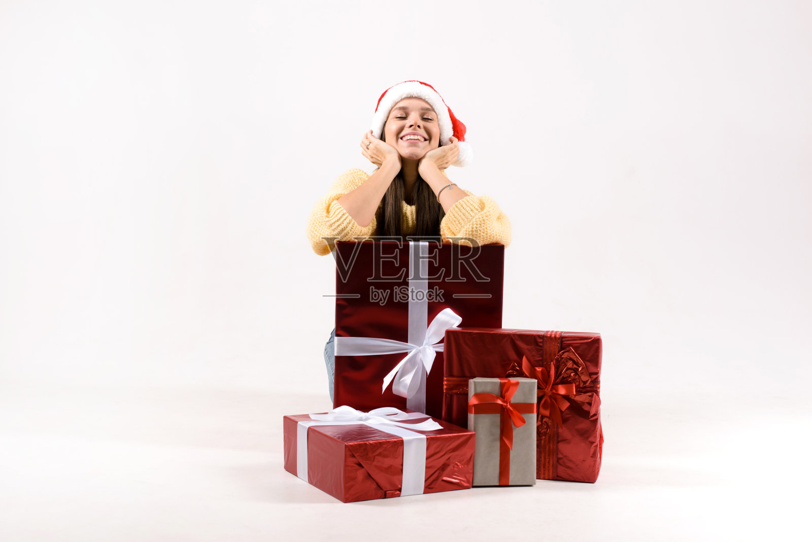 快乐的漂亮女人在红色的圣诞老人帽呆在靠近礼物圣诞盒子的堆栈孤立在灰色的背景照片摄影图片