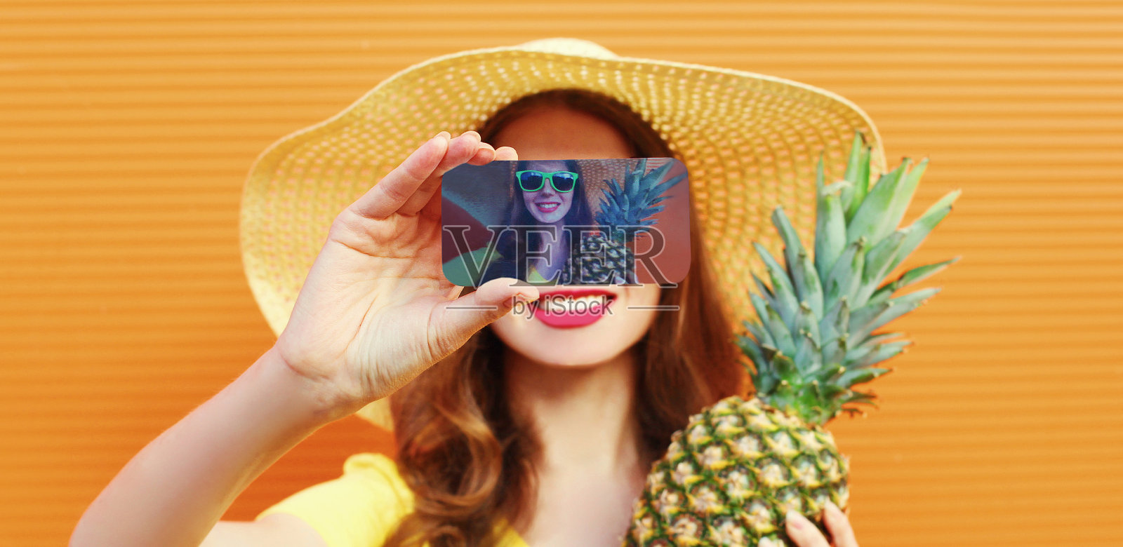 近距离的女人伸展她的手通过手机与菠萝在夏季草帽在一个橙色的背景自拍照片照片摄影图片