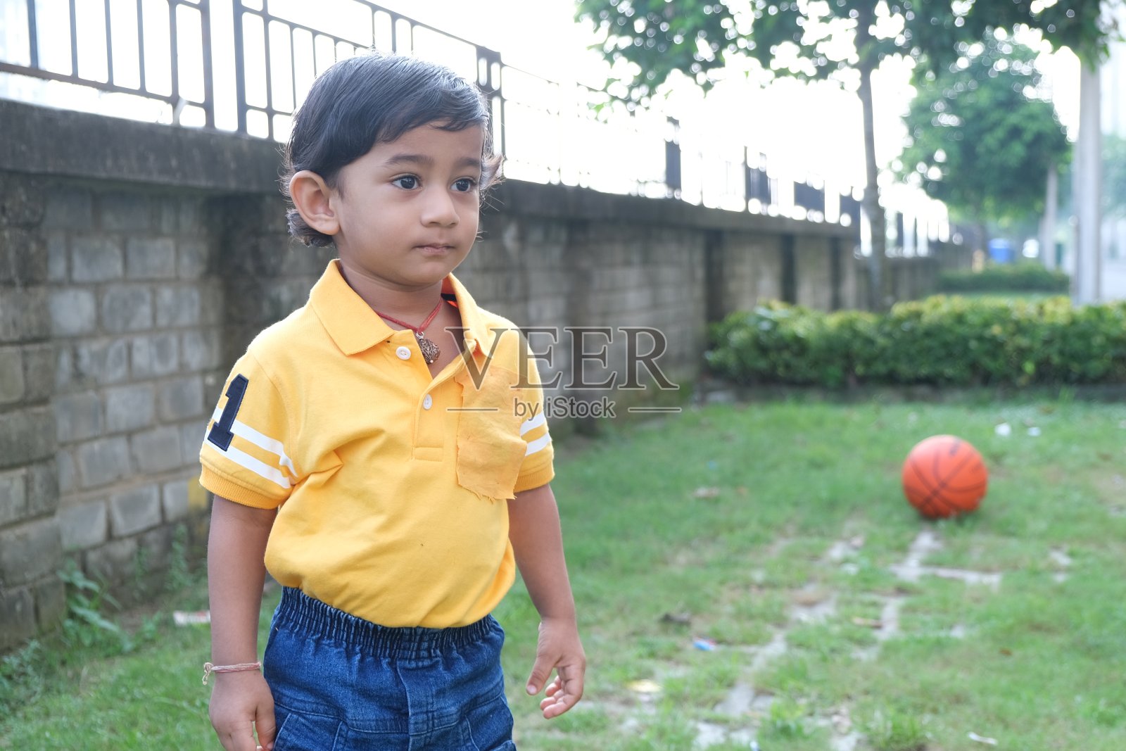 印度可爱的小愉快的深褐色的泰米尔婴儿男孩在充满活力的黄色休闲服玩自己在绿色自然背景的一个橙色球。照片摄影图片