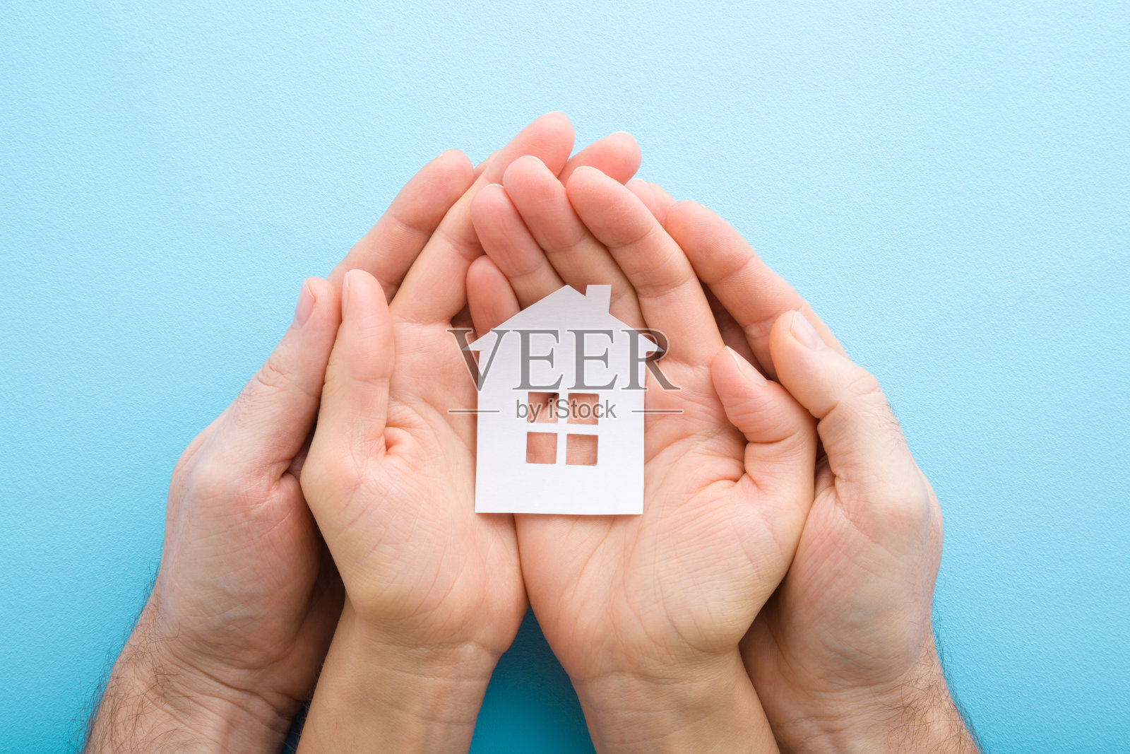 白色的纸房子在年轻的成年妇女和男子的手掌上浅蓝色的桌子背景。柔和的颜色。特写镜头。视角拍摄。自顶向下的观点。照片摄影图片
