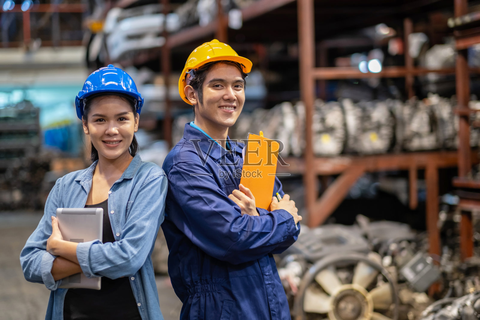 工程师们戴着安全帽，双手交叉站在汽车零部件仓库里。维护和服务理念。男人拿着写字板，女人拿着写字板。照片摄影图片
