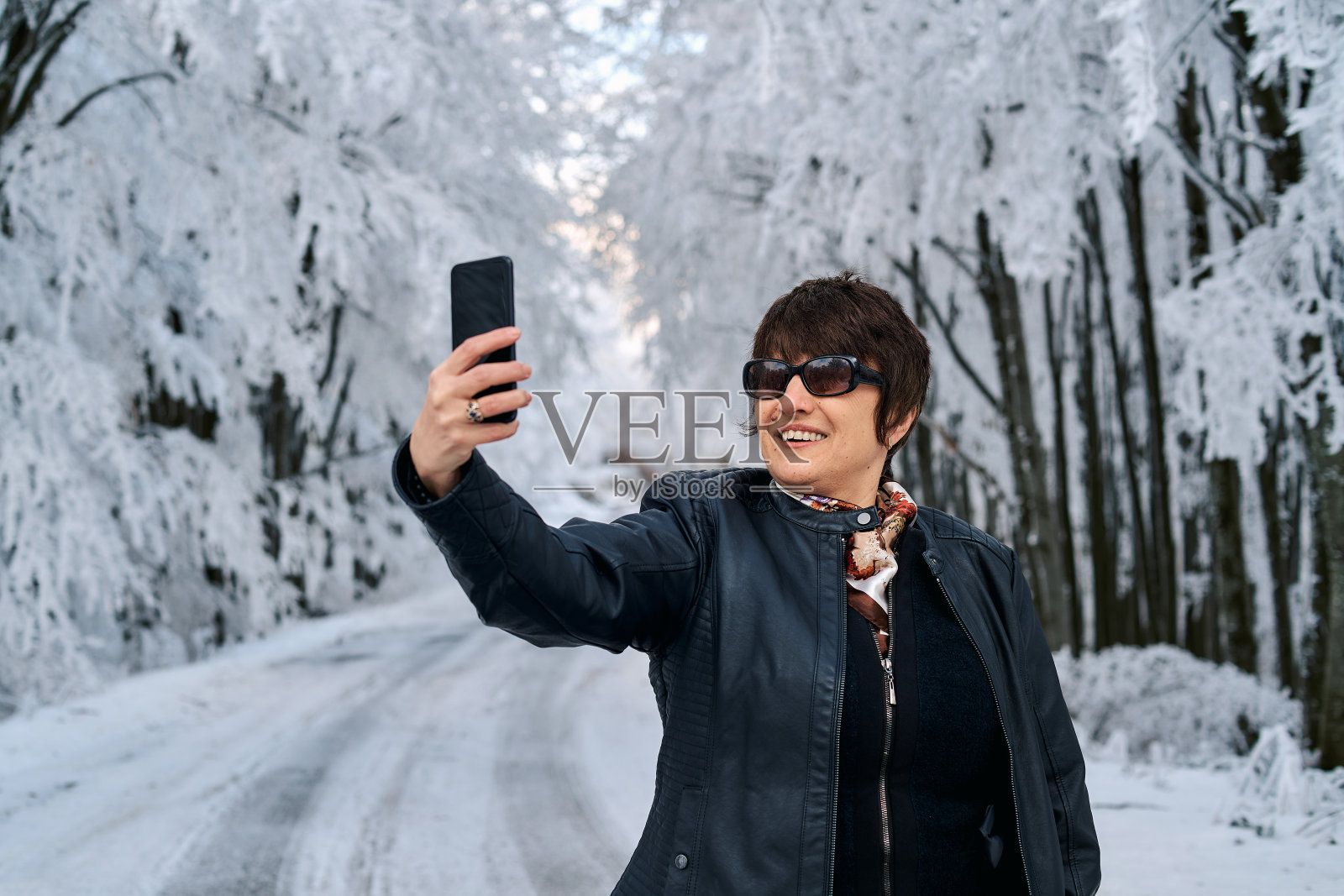 快乐微笑的成熟女人在冬天的森林里用智能手机自拍。照片摄影图片