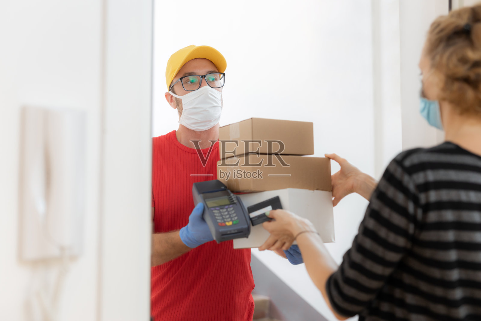带医用口罩的快递员和在病毒大流行期间在家用非接触式信用卡付款的妇女。照片摄影图片