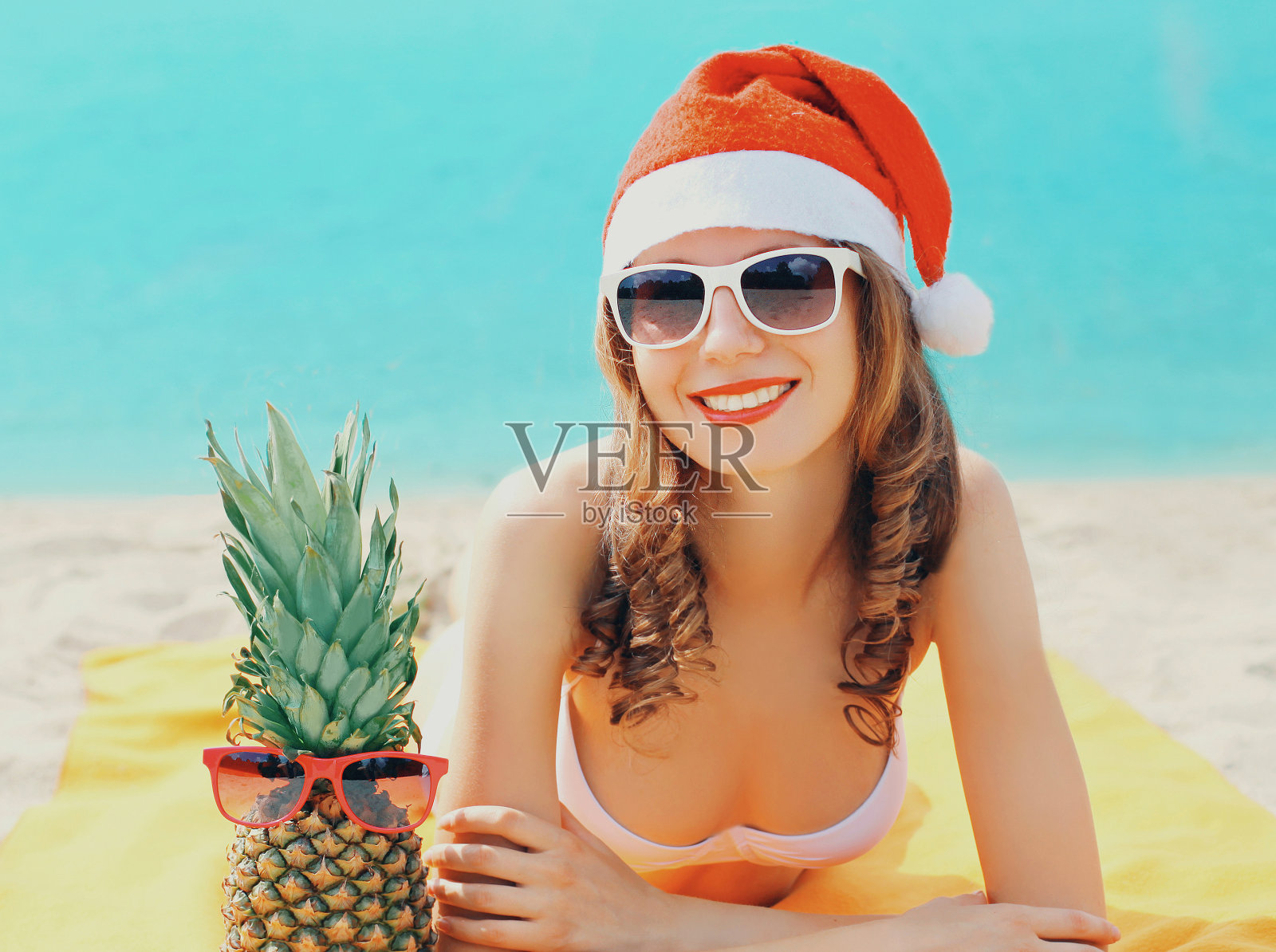 圣诞节快乐的年轻微笑的女人在红色圣诞老人帽与有趣的菠萝躺在一起在蓝色的海洋背景的海滩上的圣诞肖像照片摄影图片