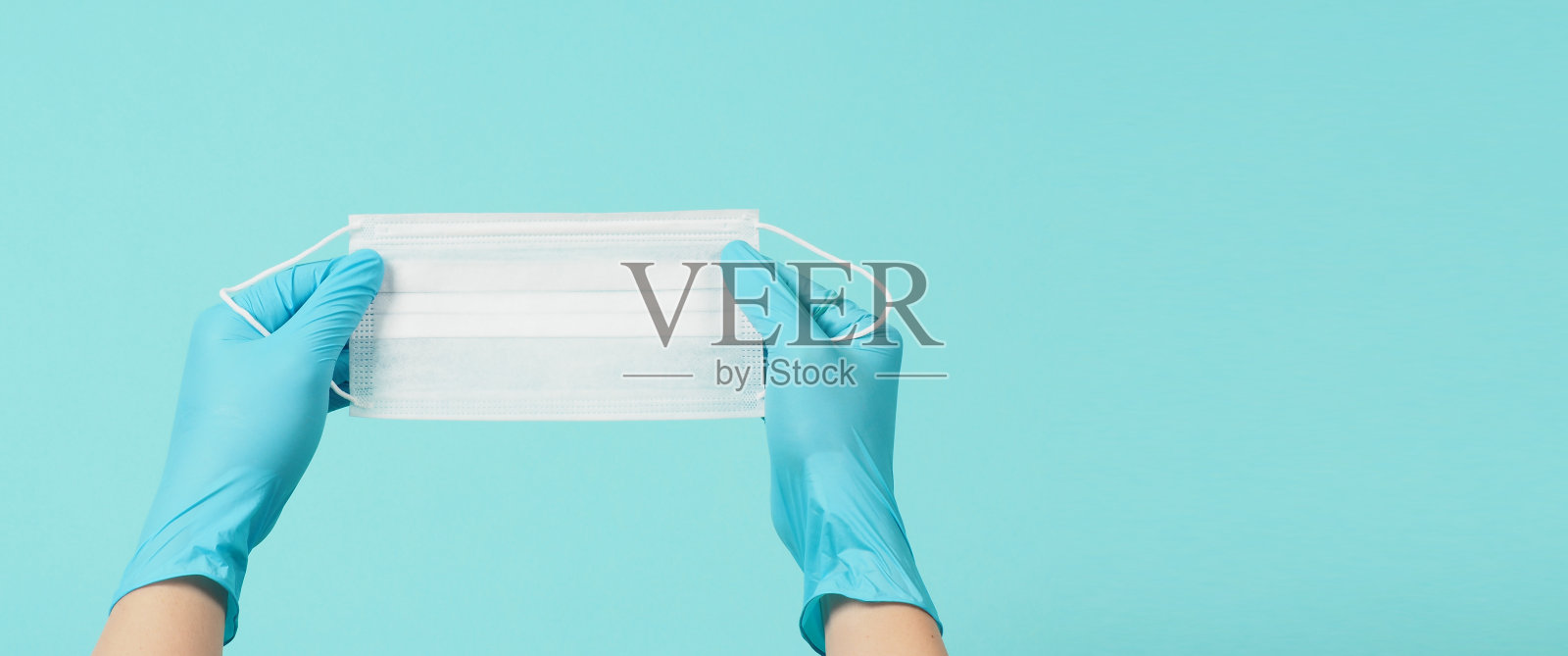 双手戴医用手套或乳胶手套，手持医用口罩，隔离在蓝色和绿色或绿松石色背景下。照片摄影图片