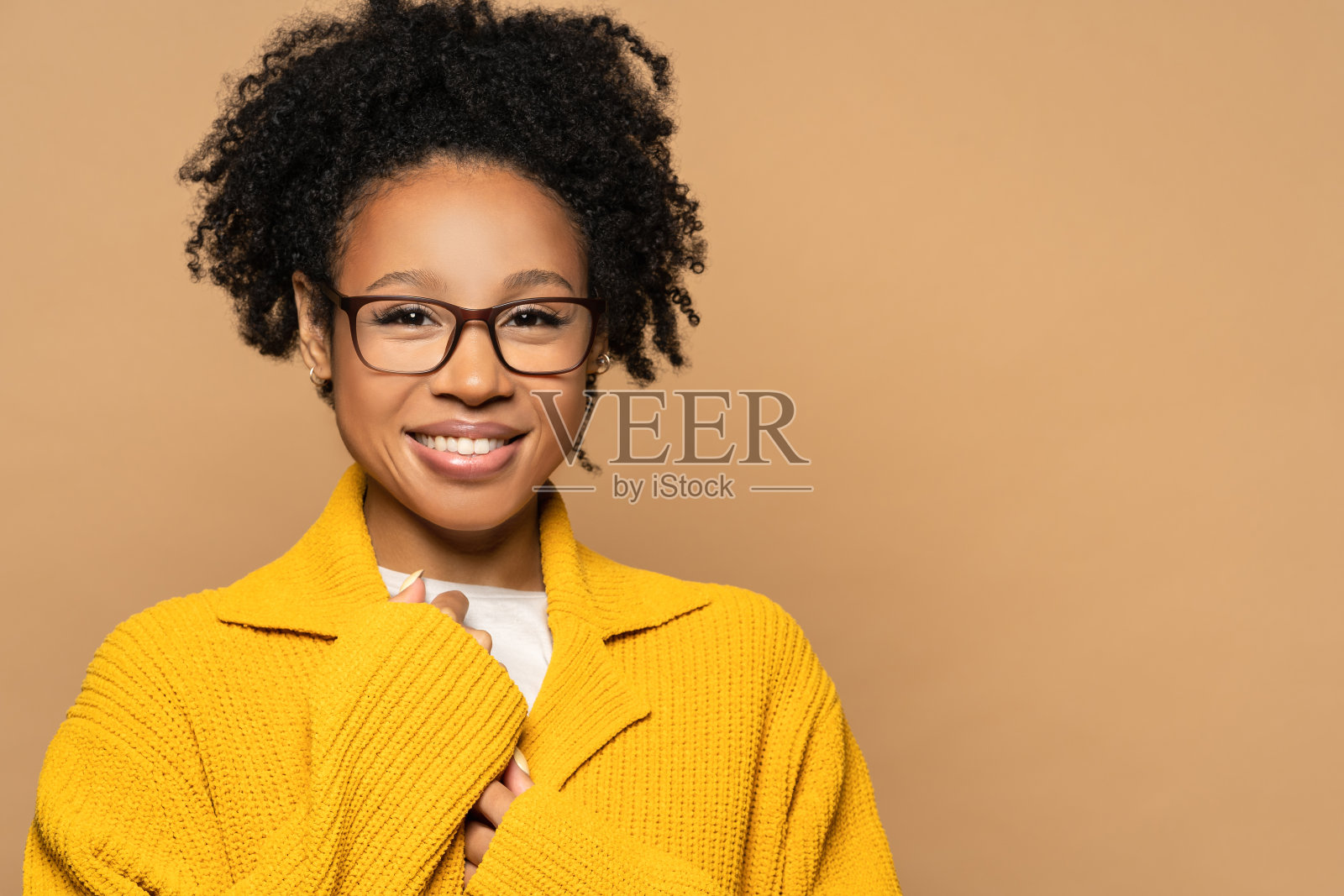 自信快乐的非洲裔美国年轻女子穿着黄色羊毛衫戴着眼镜，看着照相机。微笑的混血女孩戴眼镜隔离在工作室米色背景。照片摄影图片
