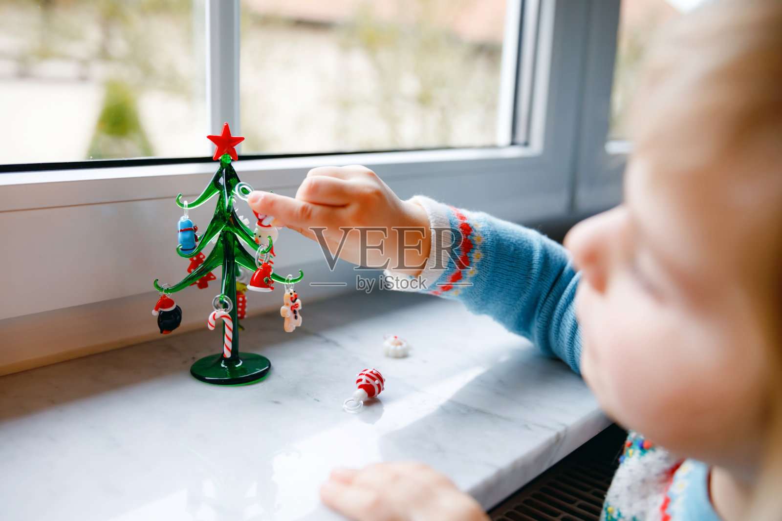 小女孩坐在窗边，用小小的圣诞玩具装饰小玻璃圣诞树。快乐健康的孩子庆祝家庭传统节日。可爱的婴儿。照片摄影图片