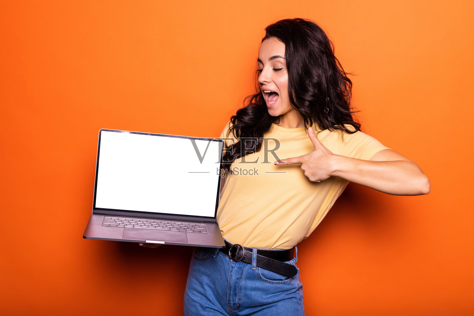 微笑年轻的黑发女人手提电脑与空白屏幕在白色t恤摆孤立的黄色背景。人们生活方式的概念。照片摄影图片