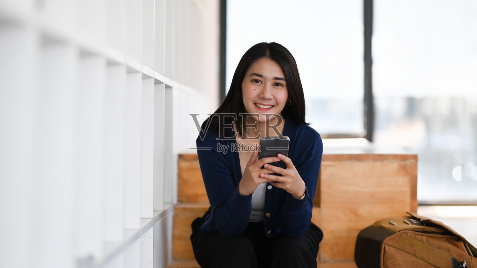 一个快乐的年轻女人手拿着智能手机，对着相机微笑，坐在图书馆里。照片摄影图片