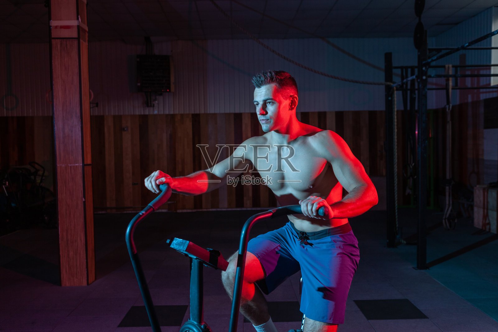 肌肉发达的男子在红蓝色霓虹灯下训练空中自行车。交叉训练照片摄影图片