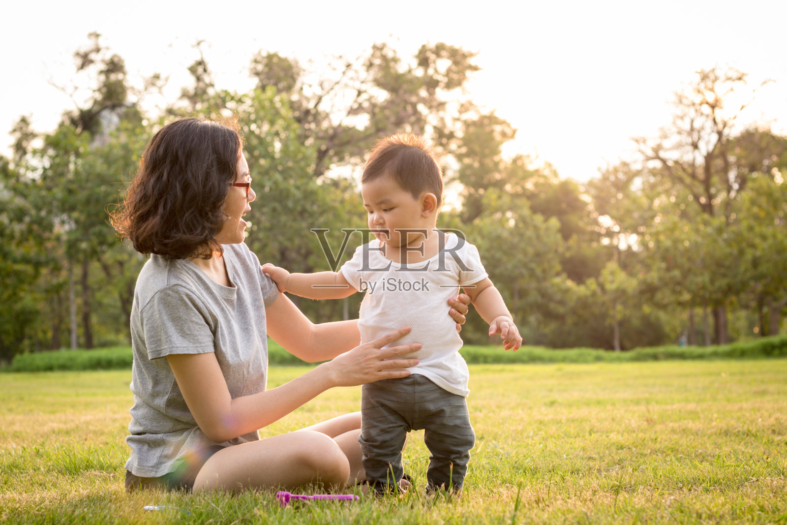 母亲帮助儿子在城市公园的草坪上学习走路照片摄影图片