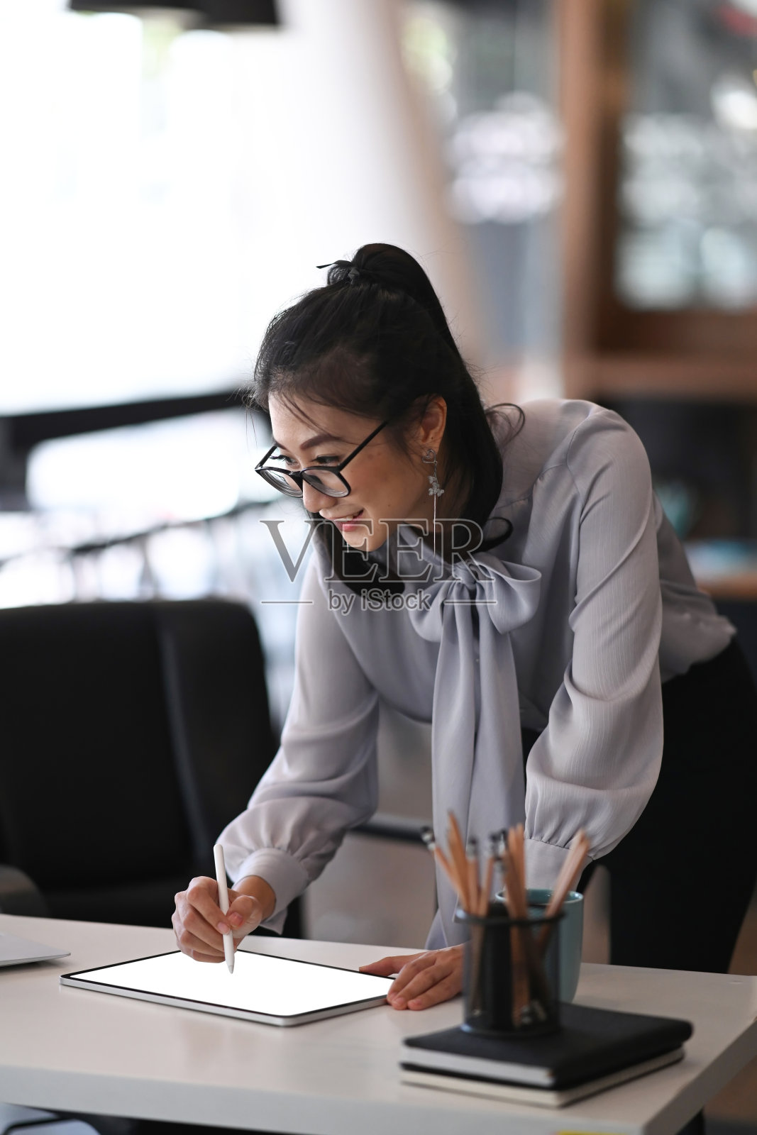 一个戴着眼镜的快乐女人的肖像站在她的工作空间和使用数字平板电脑。照片摄影图片