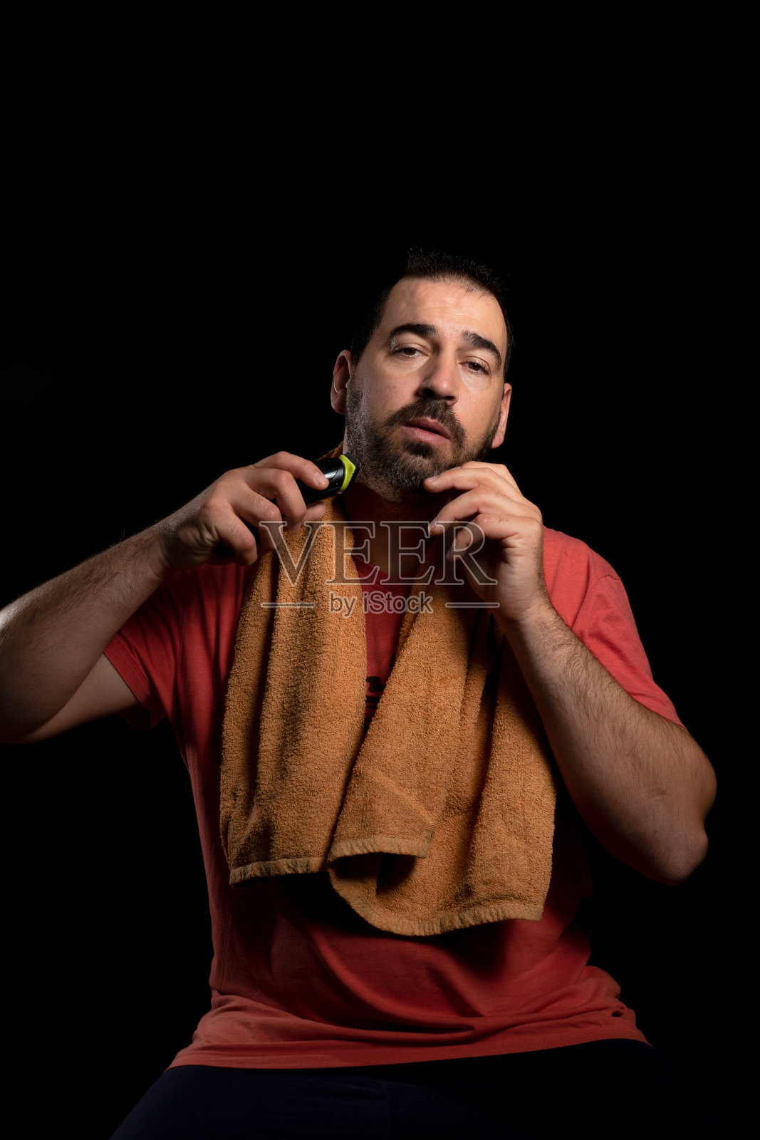 胡子男子准备用电动剃须刀刮胡子，棕色毛巾挂在脖子上，孤立在黑色背景中照片摄影图片