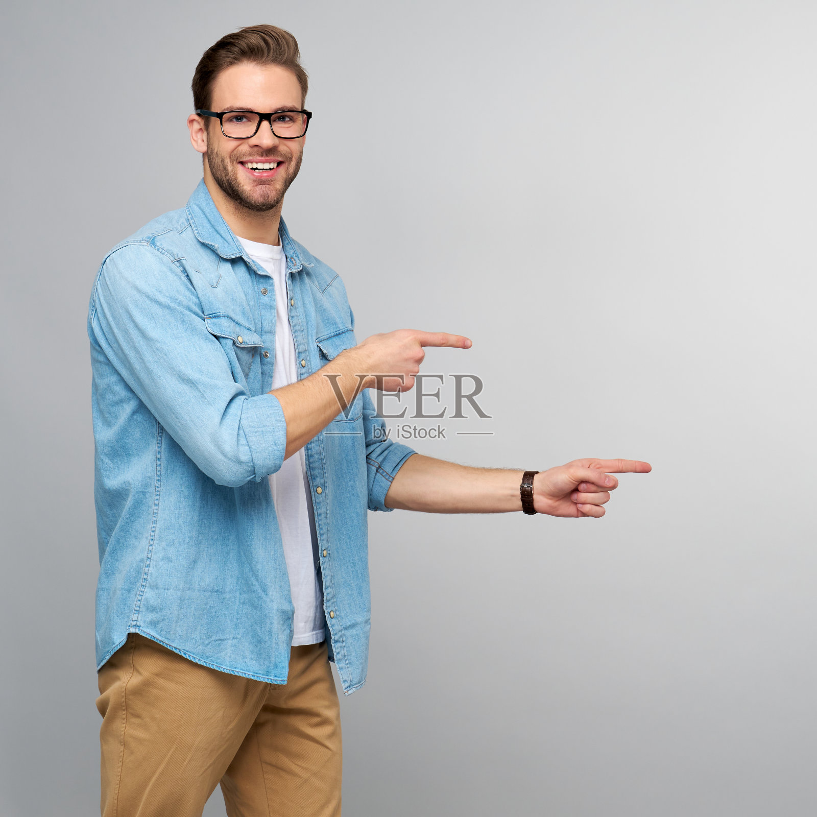 快乐的年轻英俊的男人在牛仔裤衬衫指向远方站在灰色的背景照片摄影图片