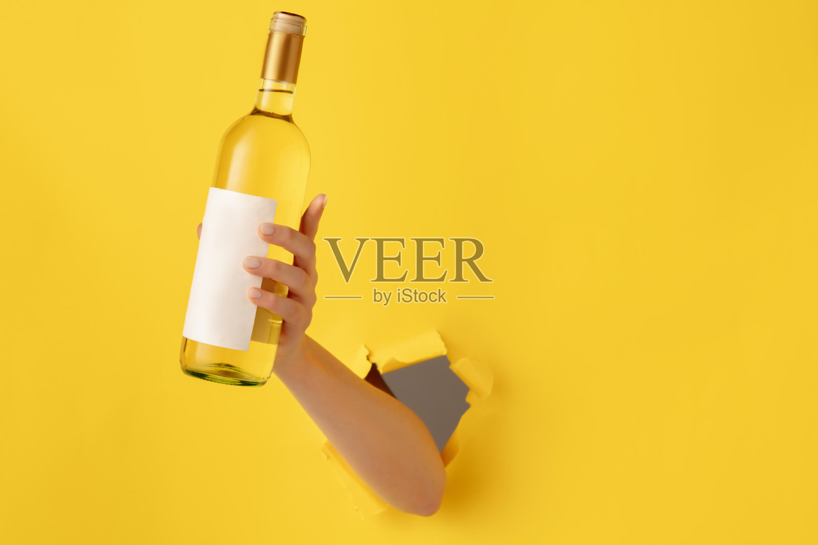 女性手拿一瓶白葡萄酒穿过黄色的纸背景。照片摄影图片