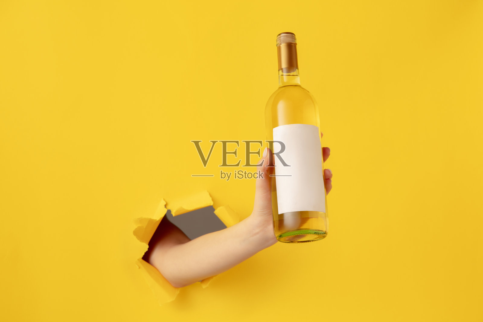 女性手拿一瓶白葡萄酒穿过黄色的纸背景。照片摄影图片