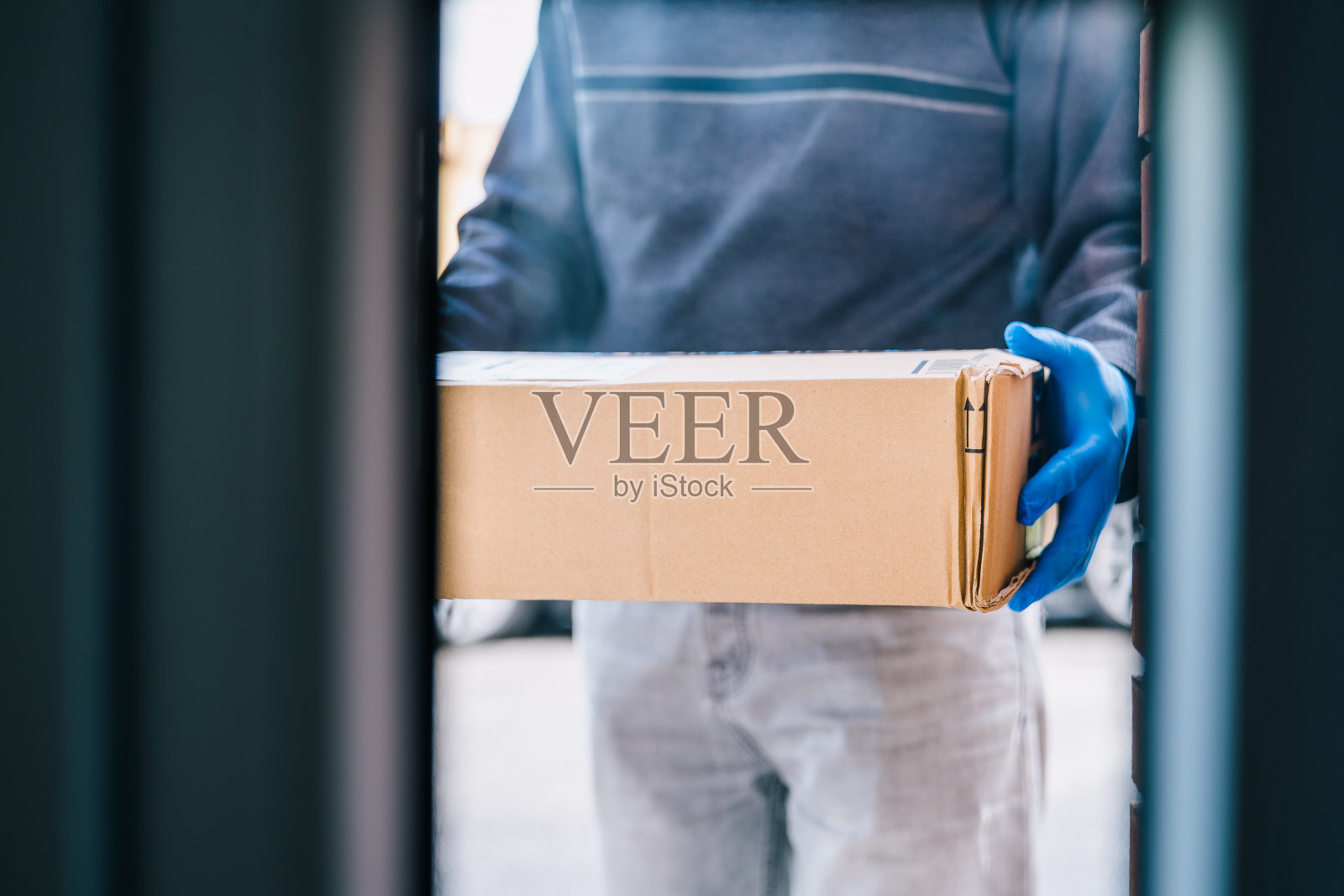 快递员将包裹留在客户门口，以防范冠状病毒感染。给一个带着蓝色乳胶手套的房子送包裹。送货员穿过玻璃门。照片摄影图片