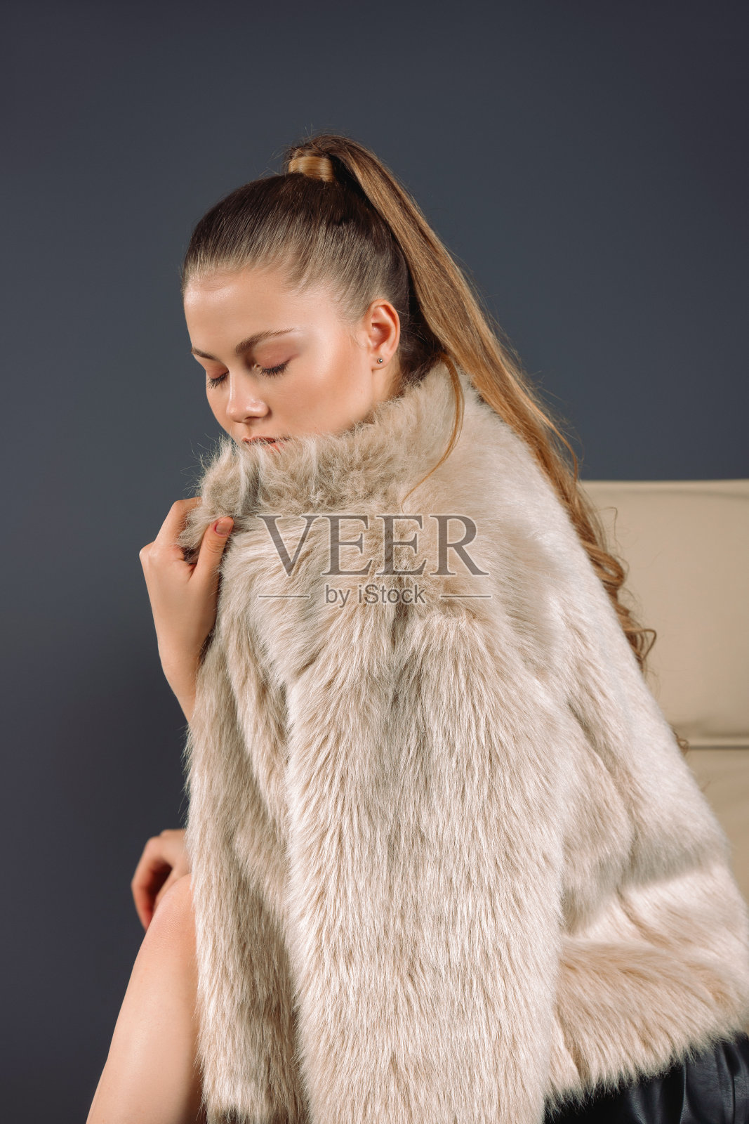 成功的女士，金发女郎坐在椅子上，穿着豪华的皮草大衣看着地板。灰色的背景。照片摄影图片