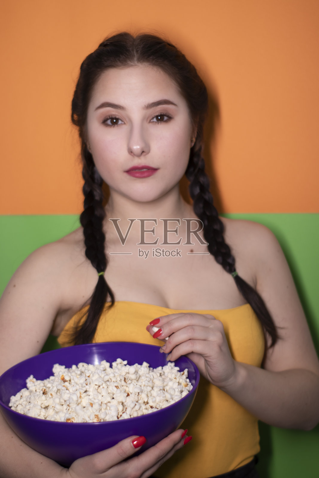 迷人的黑发女子在黄色上衣拿着一个紫色的碗爆米花在明亮的绿色和橙色的背景。电影的概念。电影照片摄影图片