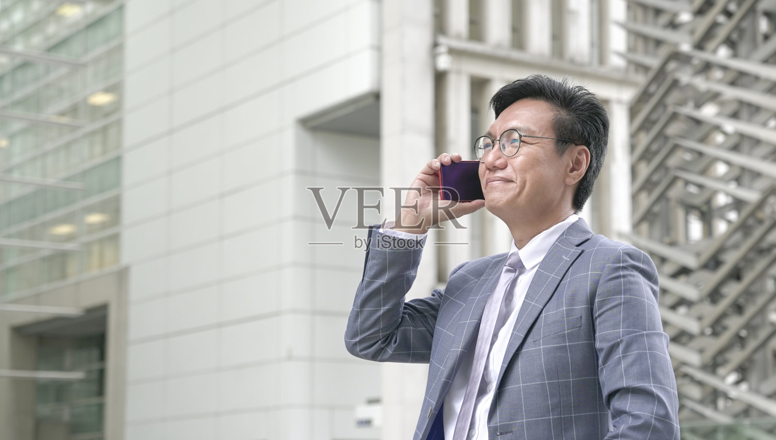 穿西装打领带的商人，微笑着讲电话，背景是现代建筑。照片摄影图片