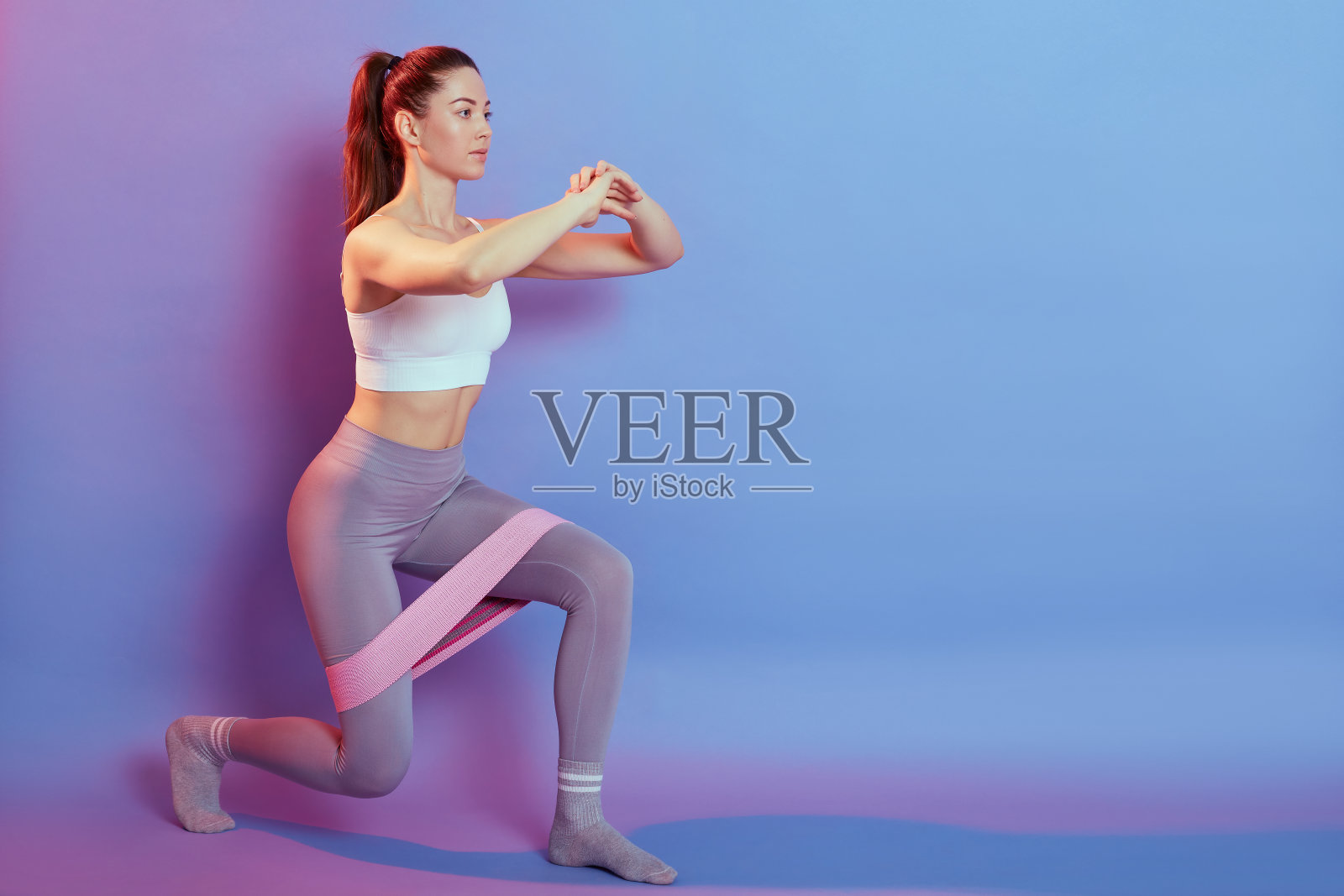 年轻漂亮的白人女性穿着运动服，扎着马尾辫，用橡皮筋做弓步，保持双手高举，训练腿和臀部，复制空间的促销。照片摄影图片