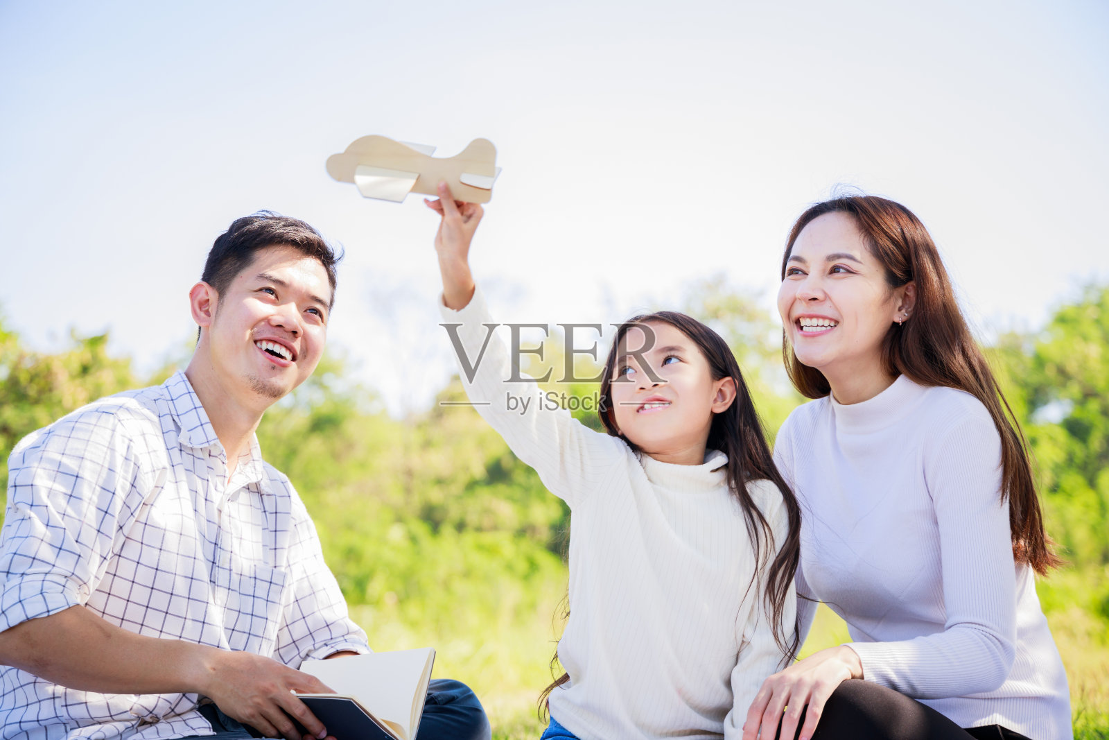 幸福的家庭野餐。亚洲父母(父亲，母亲)和女儿玩玩具飞机，在绿色的花园野餐布上享受我们自己在阳光明媚的一天照片摄影图片