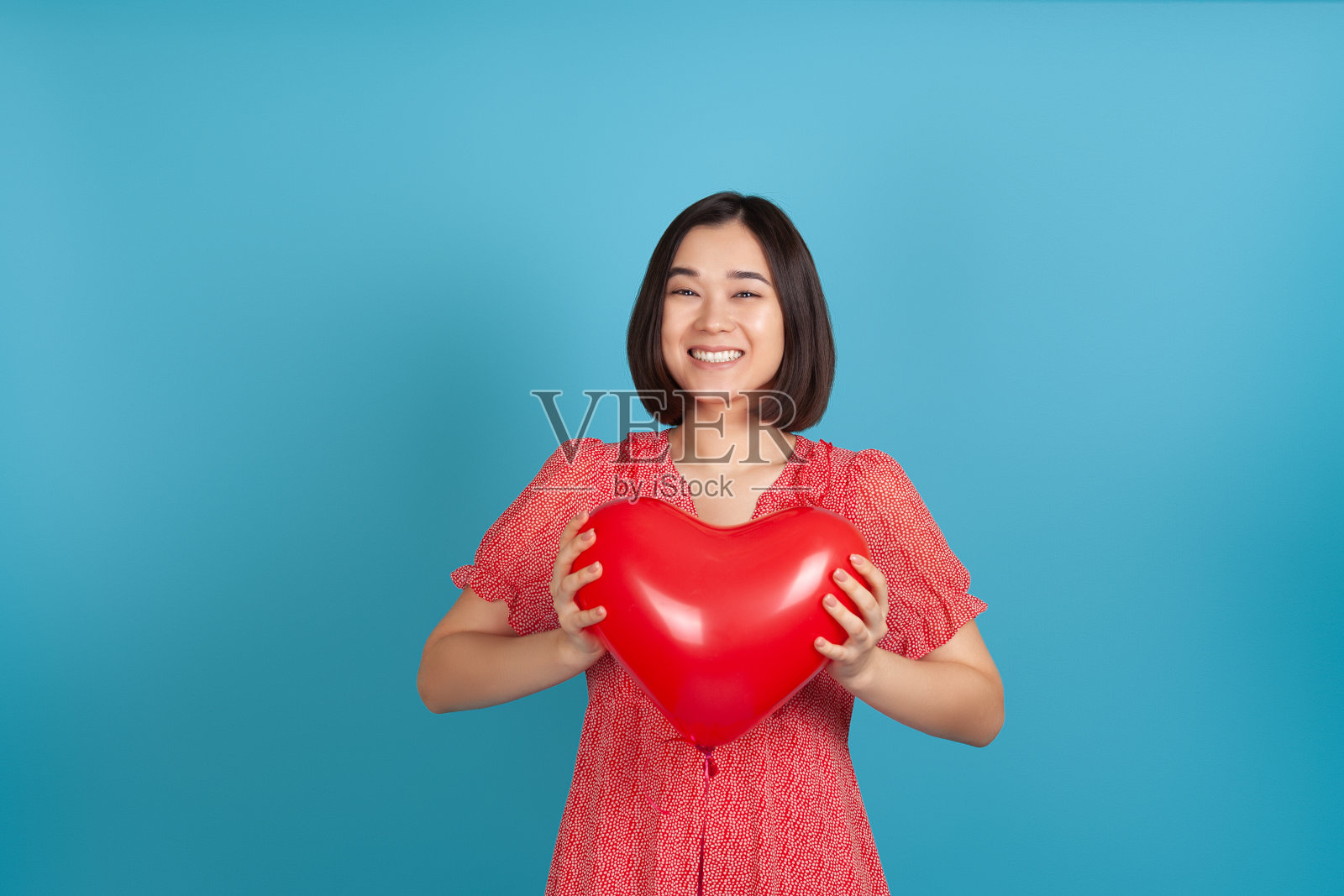 假笑满意的亚洲女人拿着红色的心形状的气球在情人节孤立在蓝色的背景照片摄影图片