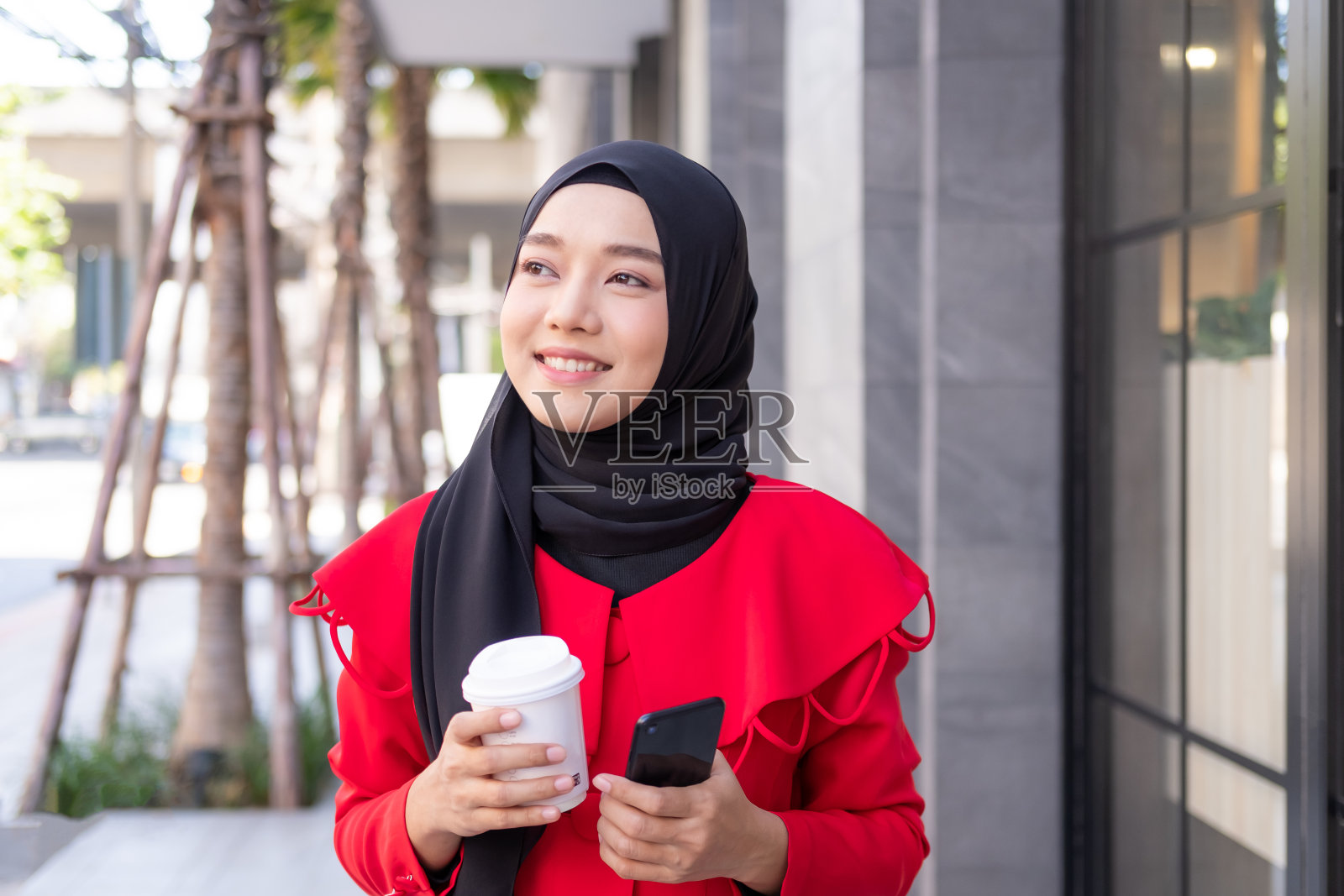 美丽的东亚伊斯兰妇女戴着头巾，穿着正式的服装。在城市地区或百货商店里拿着咖啡杯边走边用手机交谈。人们的生活方式概念。照片摄影图片