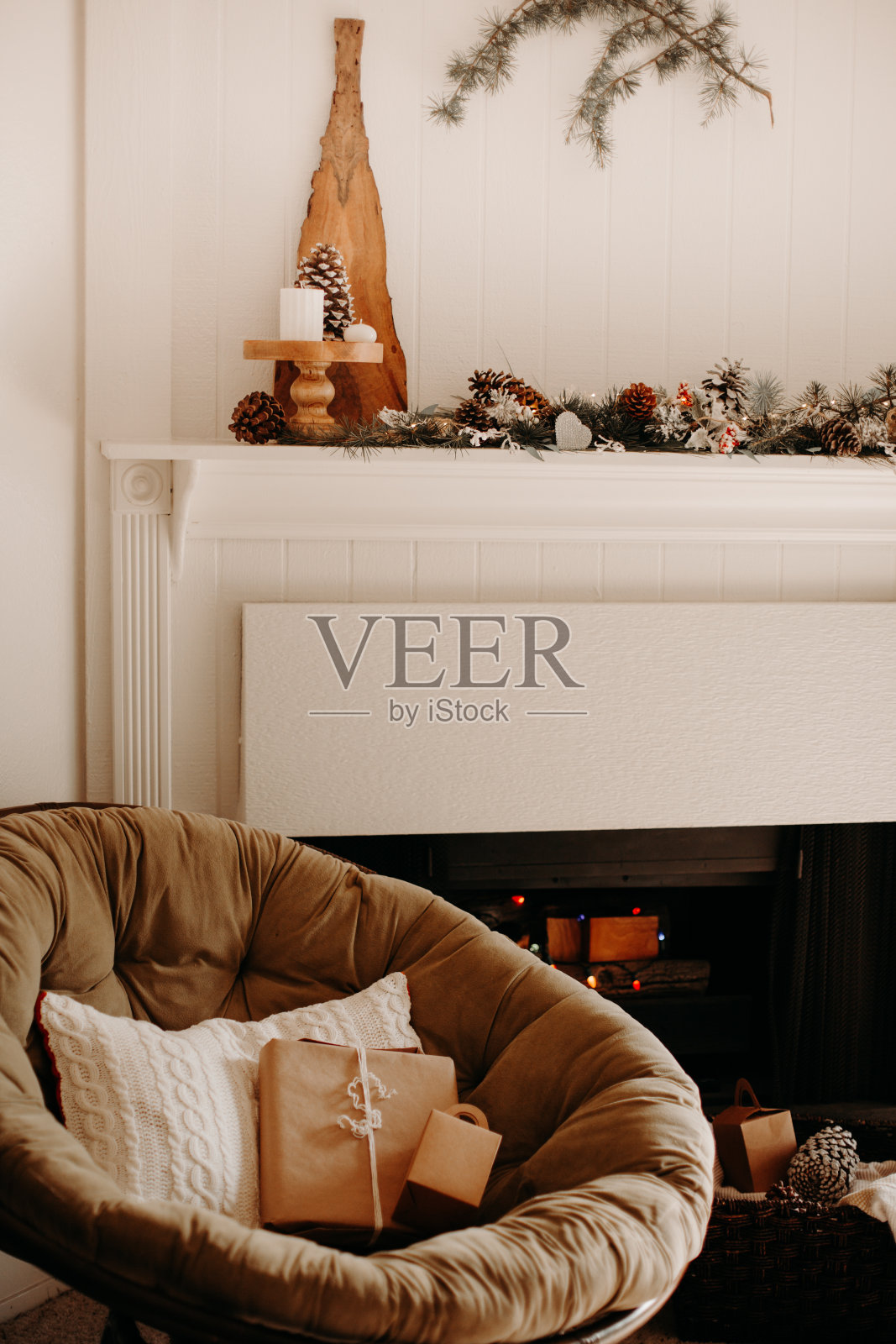 客厅和壁炉的冬季装饰——松果和圣诞树的树枝，串着灯和雪的花环，蜡烛，舒适的椅子和编织的棕色纸礼物和枕头照片摄影图片