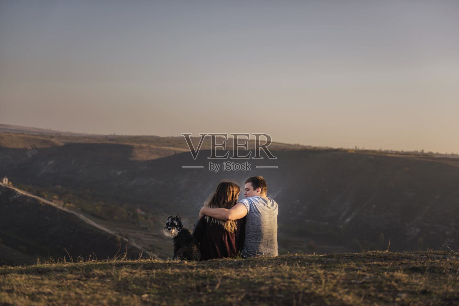 一个年轻人拥抱一个漂亮的女孩，一只雪纳瑞狗坐在旁边。幸福的家庭坐在山顶上，看着一个有河流和山脉的山谷。周末旅游，宠物旅游照片摄影图片