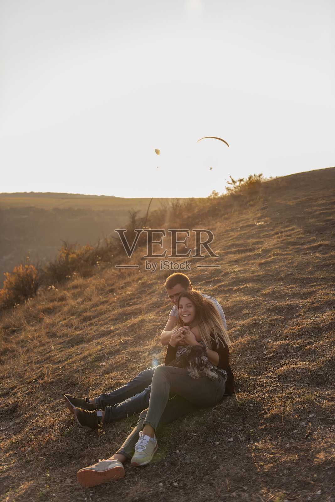 一个年轻人从后面拥抱一个美丽的女孩，两人在山顶上欣赏日落。这对恋人坐着，抱着那只小雪纳瑞。一个幸福的家庭和一只狗在秋天旅行照片摄影图片