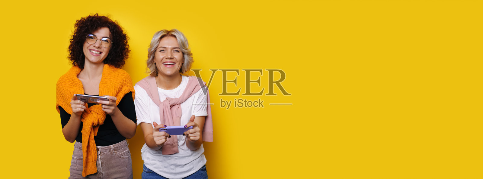 戴着眼镜的卷发女人正在玩手机，同时在工作室的黄色墙上为一款新游戏做广告照片摄影图片