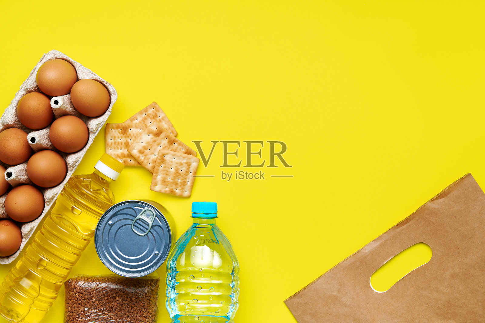 背景黄色有食品供应的面包屑、饼干、荞麦、鸡蛋、塑料瓶水、罐装食品、葵花籽油的检疫期纸袋。Copyspace。Flatlay照片摄影图片