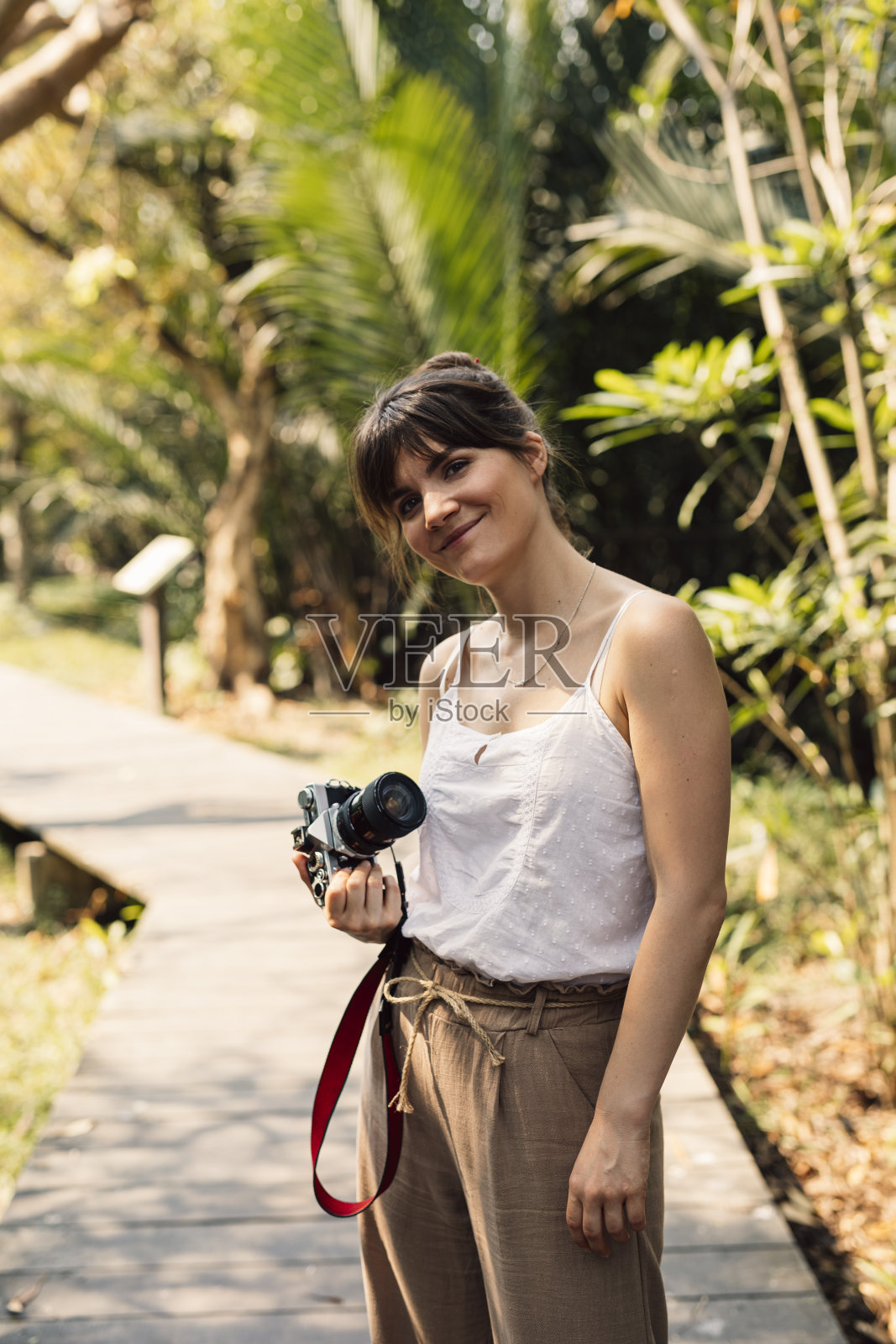 美丽快乐的女商人拿着一个复古风格的相机站在一个热带公园的木路上照片摄影图片