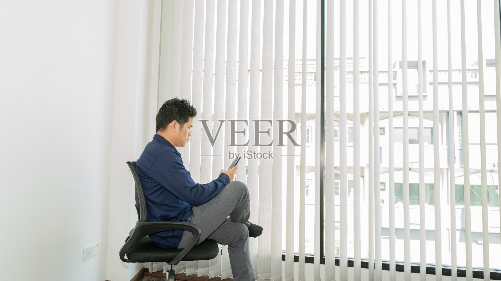 一个穿着蓝色衬衫的年轻人正坐在椅子上玩手机。照片摄影图片