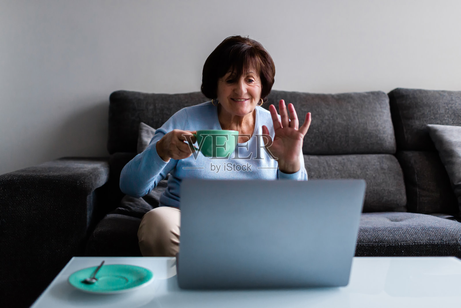 妇女坐在家里的沙发上，一边喝咖啡，一边用笔记本电脑开视频会议照片摄影图片