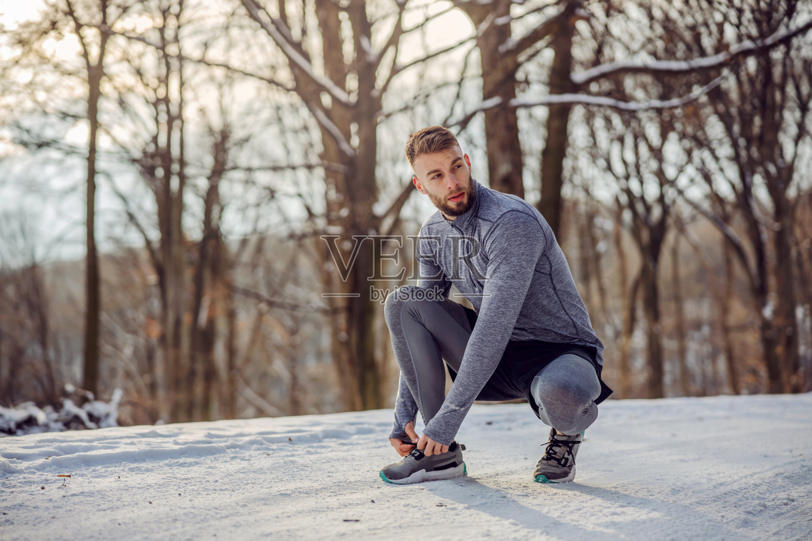 在雪天里，跑步者蹲在大自然的小路上系鞋带。健康的生活方式，运动服，下雪的天气照片摄影图片