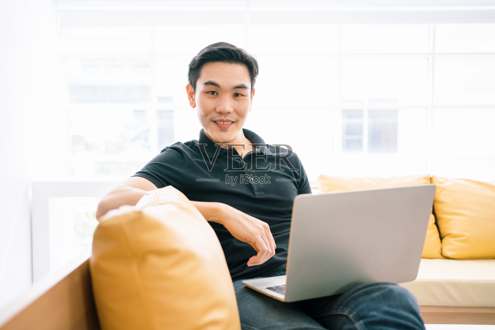 亚洲男性微笑着，一边用笔记本电脑工作，一边坐在沙发上看镜头。在家里用笔记本电脑工作的普通商人。解决业务中的问题流程。照片摄影图片