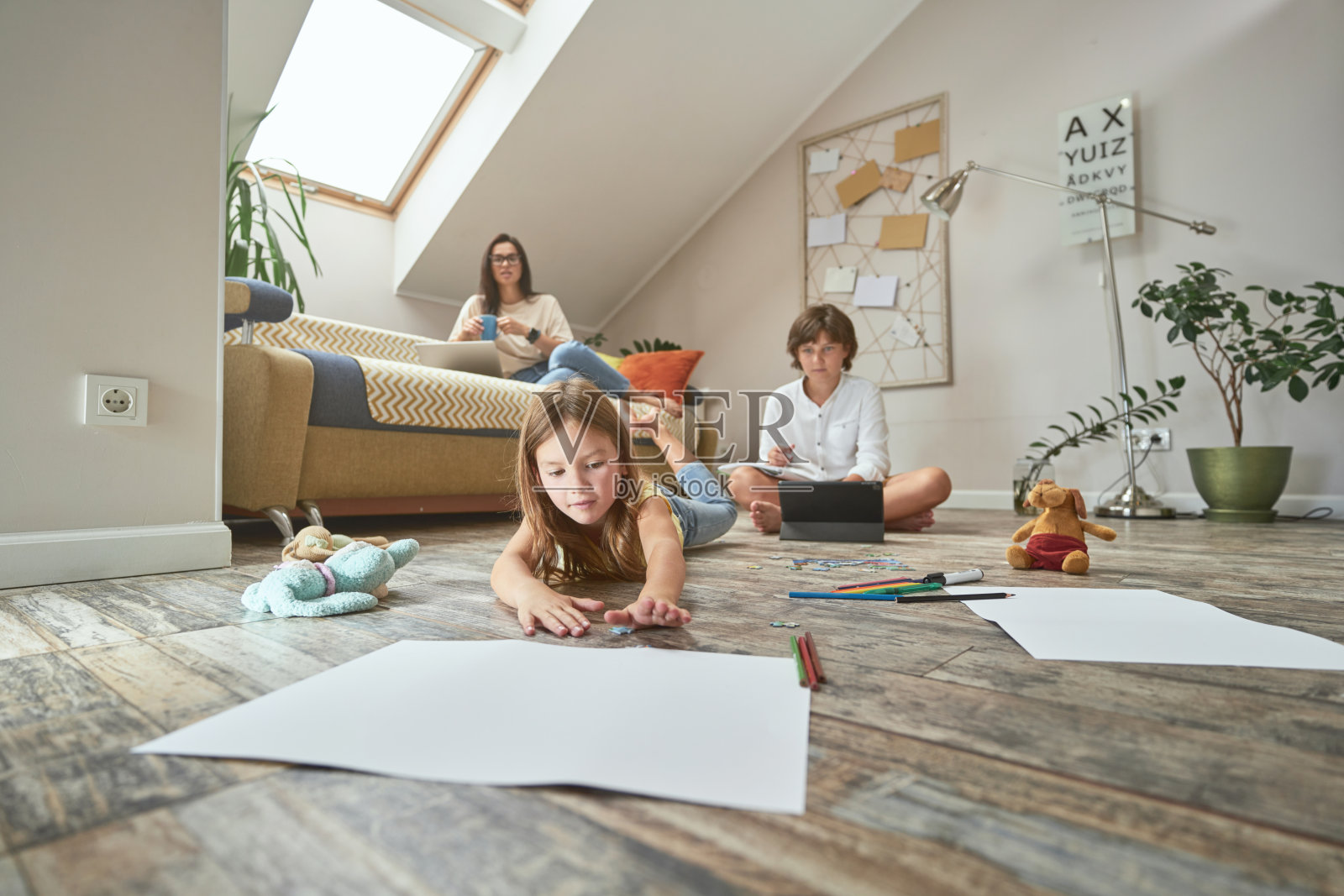 学前班的女孩躺在地板上用彩色铅笔画画，她的弟弟在网上做作业，妈妈坐在沙发上喝着咖啡，用着笔记本电脑照片摄影图片