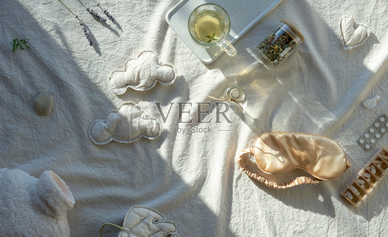 健康的夜间睡眠创意概念。浅白色，象牙色的纺织品背景。蚕丝睡眠面膜，茶，茶叶玻璃罐，睡眠软云和暖瓶。干薰衣草花照片摄影图片