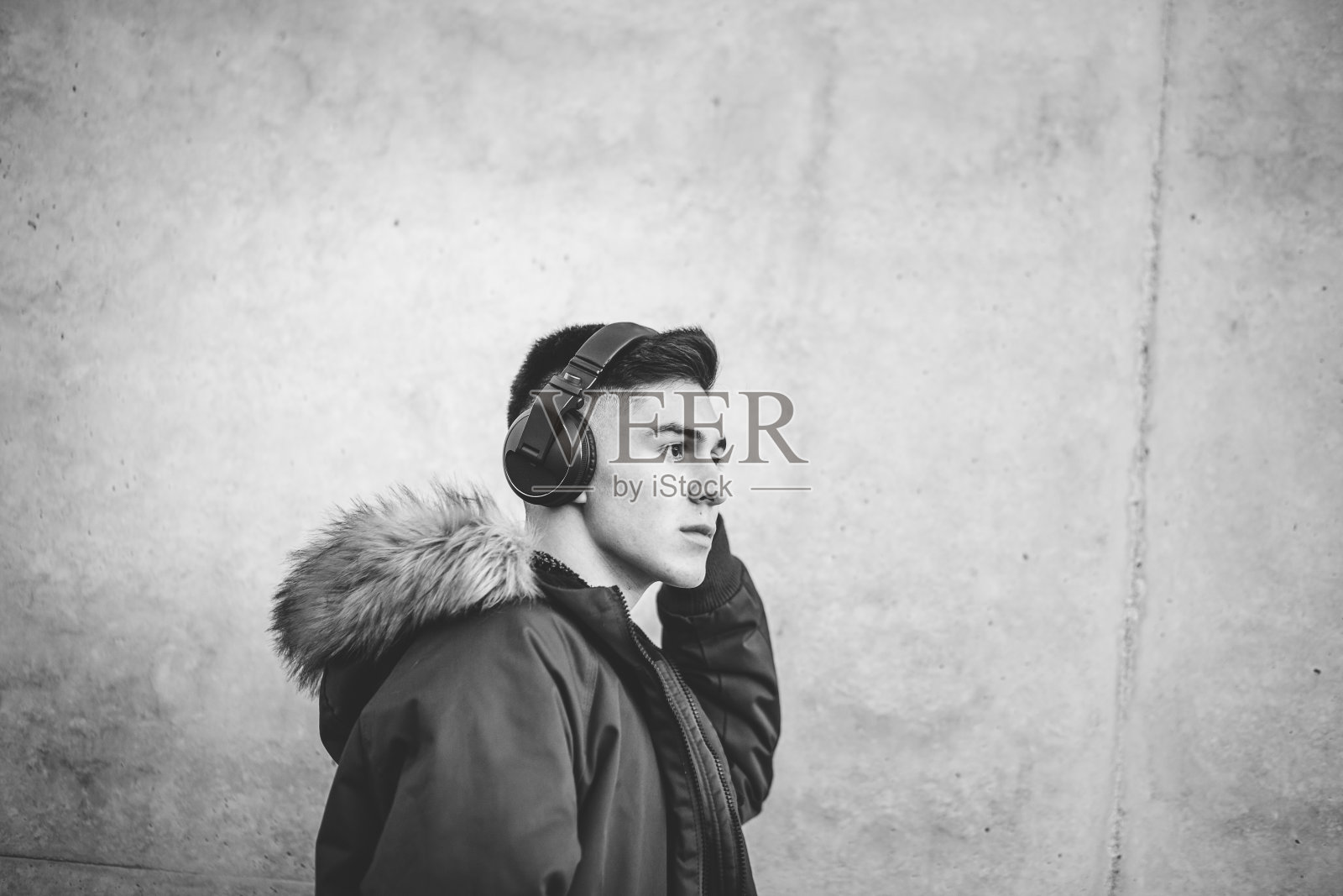 穿着夹克的年轻人戴着耳机听音乐，在混凝土背景下摆姿势。照片摄影图片