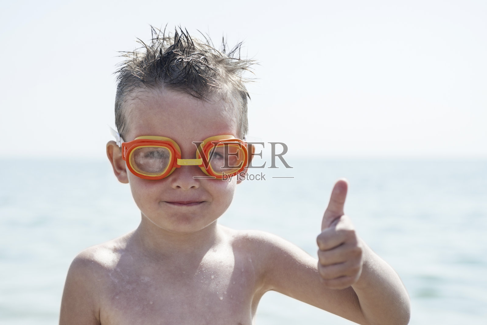 一个戴着游泳眼镜的男孩在大海的背景上竖起大拇指。戴眼镜的孩子在海里游泳照片摄影图片