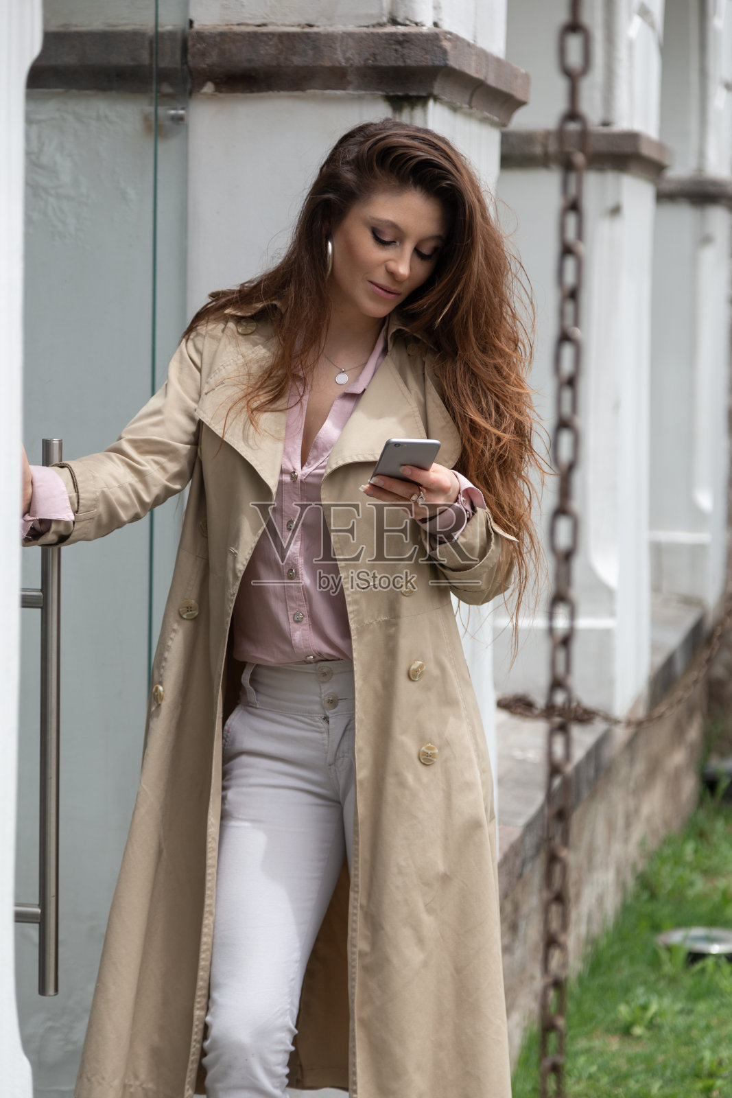年轻的拉丁女子，一头长卷发，面带微笑，手里拿着手机，穿着米色外套，粉色衬衫和白色裤子，优雅的风格在一天照片摄影图片