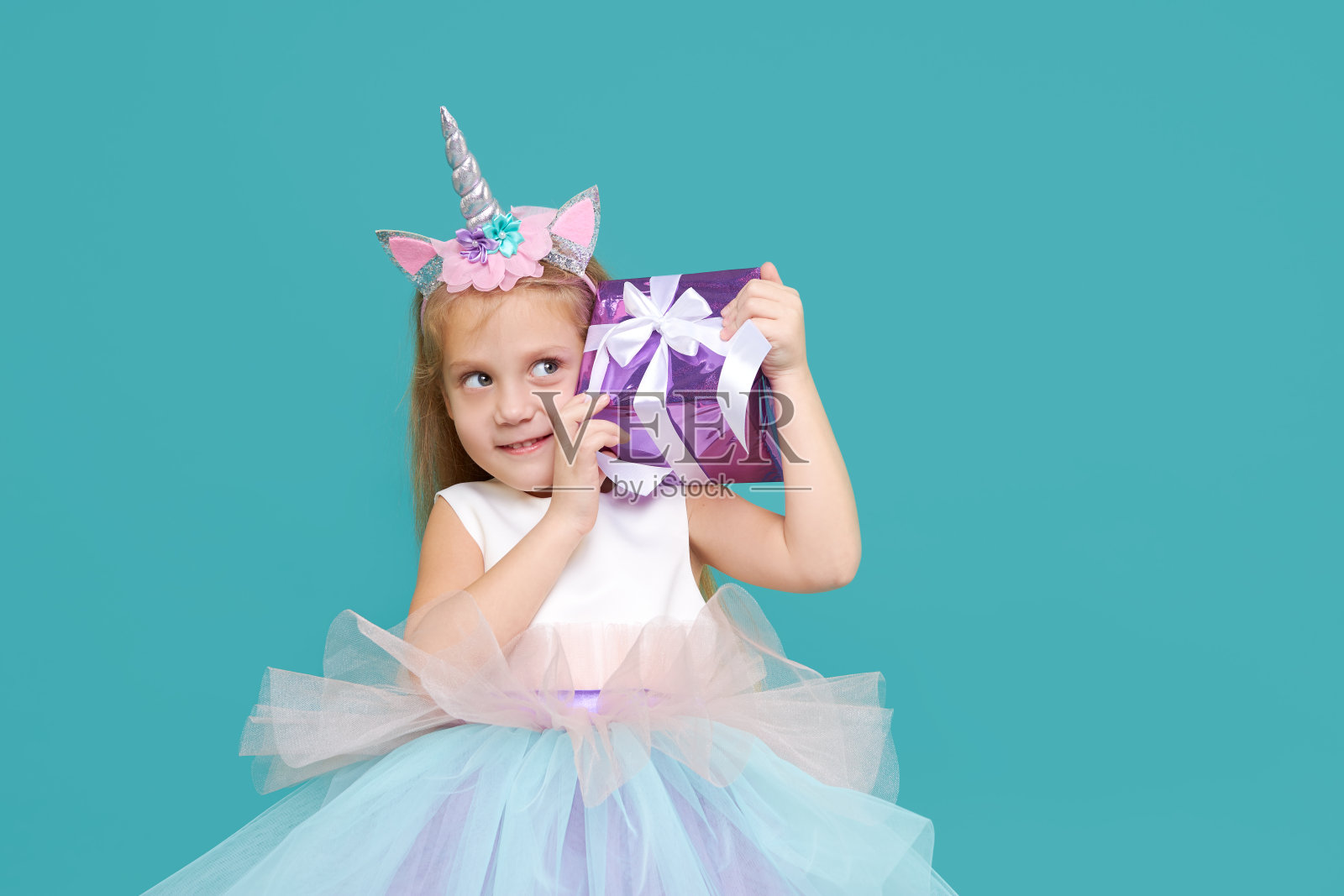可爱的小女孩在优雅的薄纱裙和独角兽头带庆祝生日派对在彩色的背景。滑稽的脸和礼盒。照片摄影图片